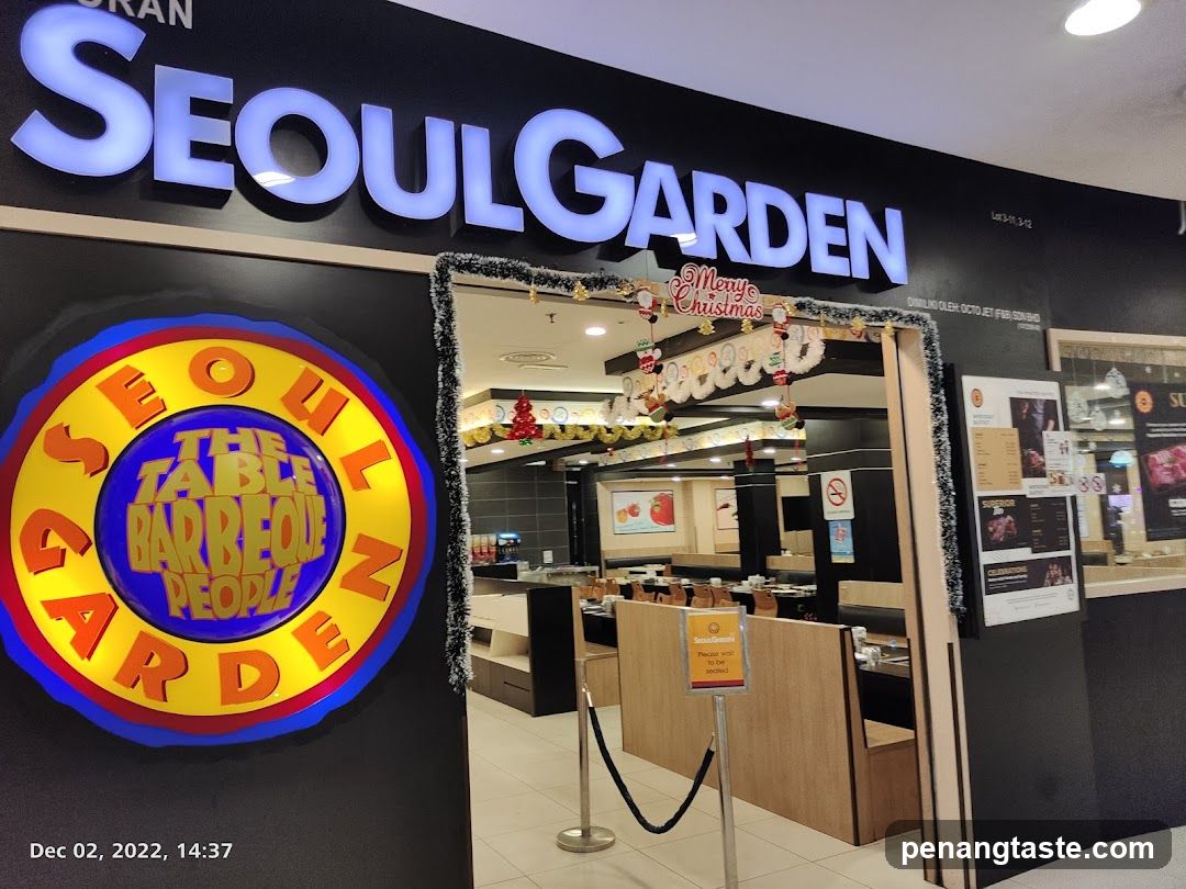 Photo of the physical storefront entrance to Seoul Garden, a Korean BBQ restaurant. The facade features large illuminated signage reading 'SEOULGARDEN' and a circular logo with 'THE TABLE BARBEQUE PEOPLE'. A festive Christmas-themed archway frames the doorway, decorated with garlands and hanging ornaments. Inside, wooden booth seating is visible along with promotional posters on the wall.